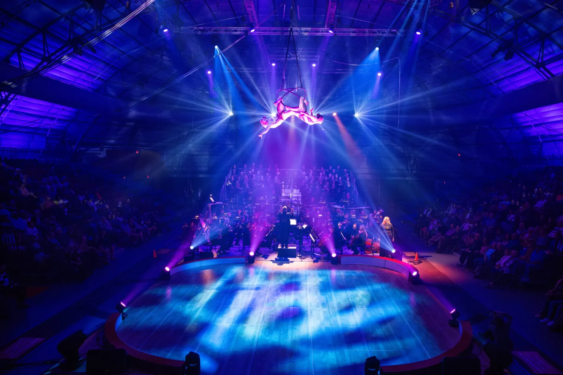 Aerial circus artists performing above the stage while Key Chorale and orchestra present a collaborative production under dramatic purple and blue lighting.
